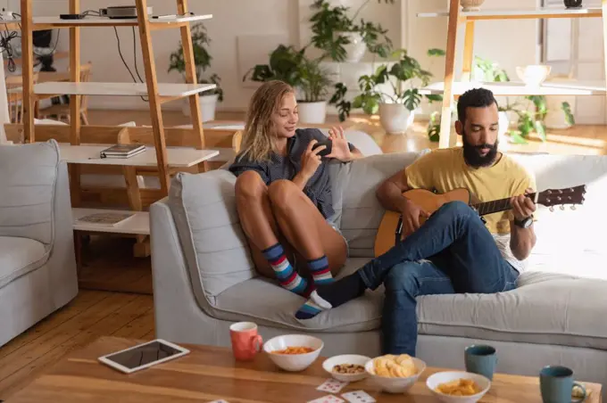 Front view of caucasian Woman using mobile phone while mixed race man playing guitar in living room at home