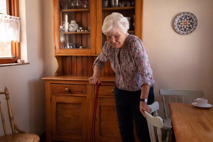 Front view of an active senior woman walking with a cane in the kitchen at home