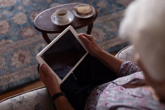 High angle view of an active senior woman using a digital tablet while sitting on the sofa at home for the teatime