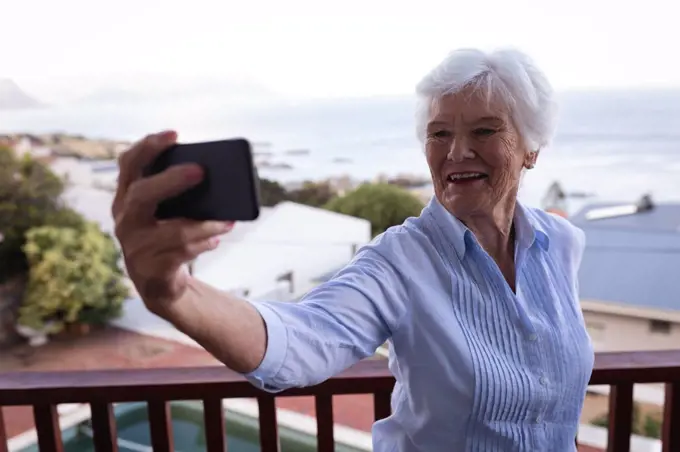 Front view of a happy active senior woman taking a selfie on the balcony against a sea landscape with her mobile phone at home