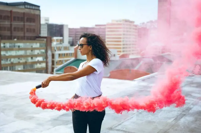 Side view of a young African-American woman wearing a white shirt and eyeglass holding a smoke maker producing red smoke on a rooftop with a view of buildings. Bright modern gym with fit healthy people working out and training