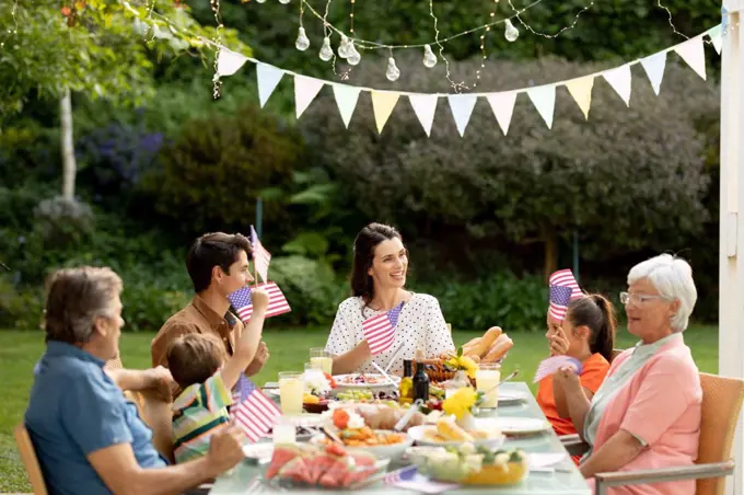 Front view of a multi-generation Caucasian family sitting outside at a dinner table set for a meal, celebrating and waving US flags. Family enjoying time at home, lifestyle concept