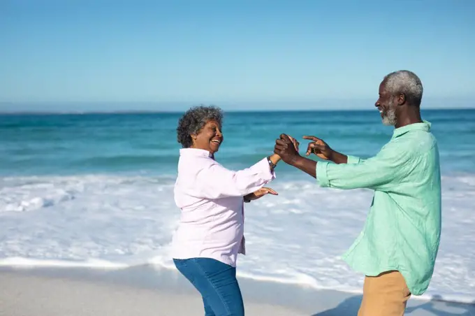 Front view of a senior African American couple standing on the beach with blue sky and sea in the background, dancing and having fun