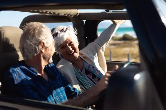 Side view close up of a senior Caucasian couple sitting in their car, driving to the beach in the sun, the man at the wheel and the woman with her hand out of the window and both smiling at each other