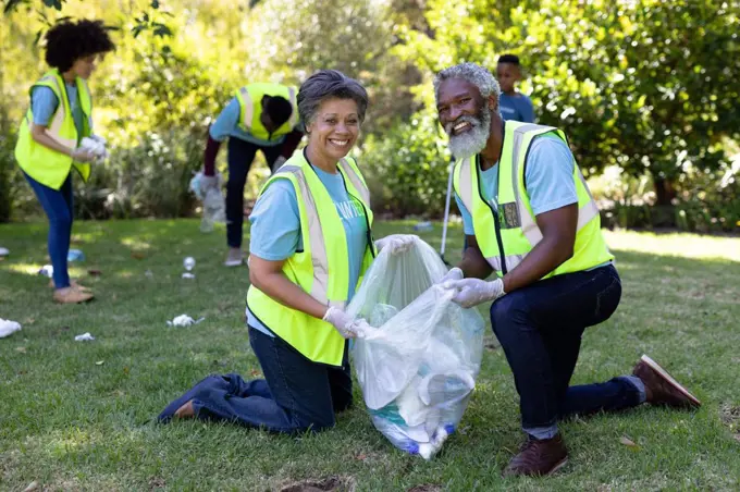 Senior mixed race couple spending time outside with their family, presenting a garbage bag with garbage in it, looking at the camera and smiling, with their family in the background, all wearing blue volunteers t shirts, on a sunny day