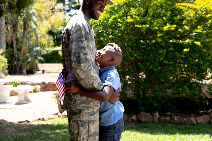 African American male solider wearing uniform embracing his son holding a USA flag, standing in their garden on a sunny day, smiling and interacting.