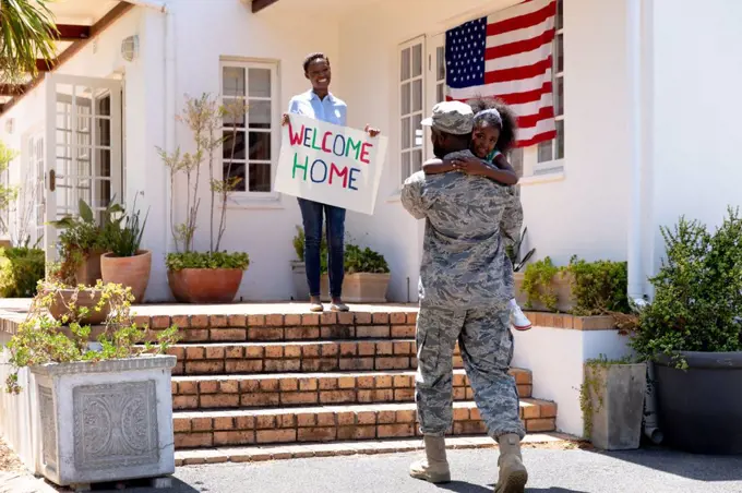 African American woman and her daughter welcoming an African American solider wearing uniform, embracing by their house on a sunny day.