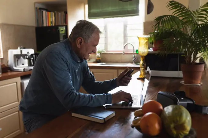 Side view of a senior Caucasian man relaxing at home, sitting at the counter in his kitchen using a smartphone and laptop computer