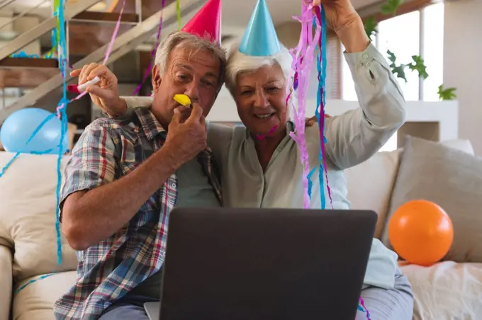 Senior caucasian couple on video call celebrating birthday. in party hats blowing party blowers. sitting in living room at home using laptop. self isolation during coronavirus covid 19 pandemic