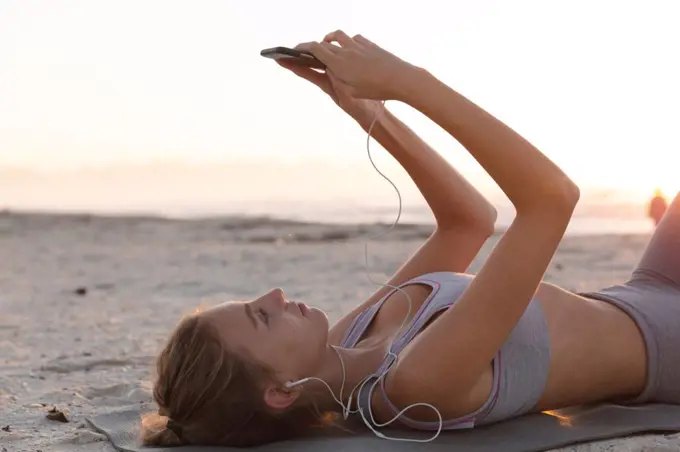 Caucasian woman wearing earphones using smartphone while lying on yoga mat at the beach. fitness yoga and healthy lifestyle concept