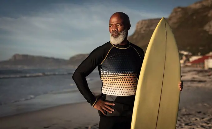 Portrait of african american senior man on beach holding surfboard looking out to sea. health and wellbeing, active retirement.