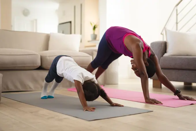Smiling african american mother and daughter practicing yoga, stretching on mats in living room. family spending time together at home.