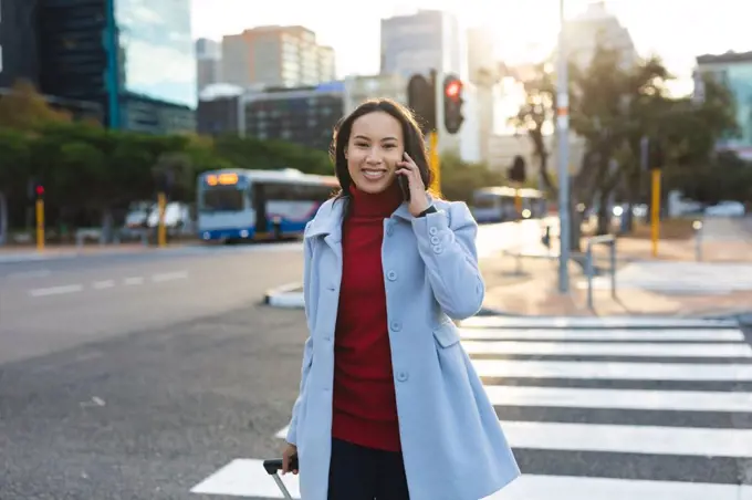 Portrait of asian woman using smartphone and crossing road with suitcase. independent young woman out and about in the city.