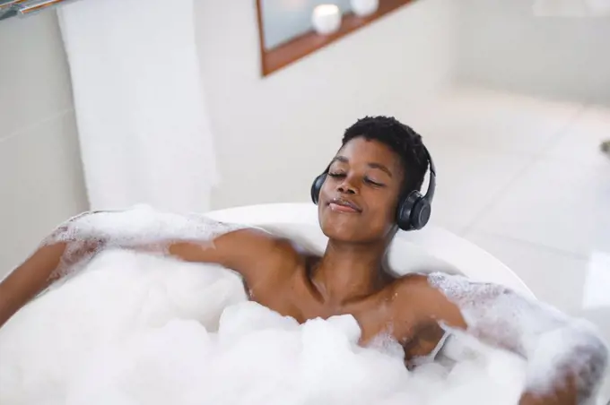 Smiling african american woman in bathroom relaxing in foam bath wearing headphones with eyes closed. domestic lifestyle, enjoying self care leisure time at home.