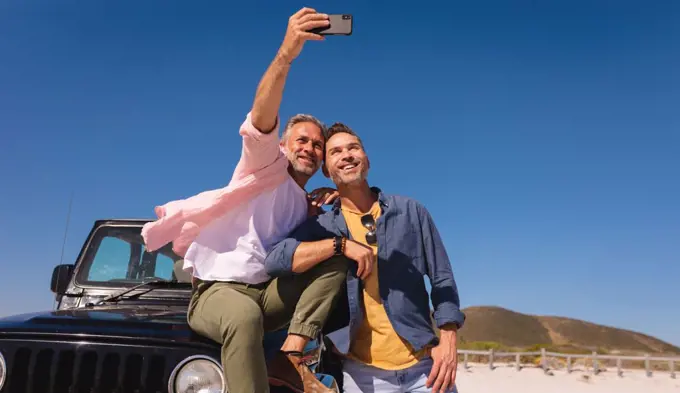 Happy caucasian gay male couple embracing and taking selfies by a car at seaside. summer road trip and holiday in nature.