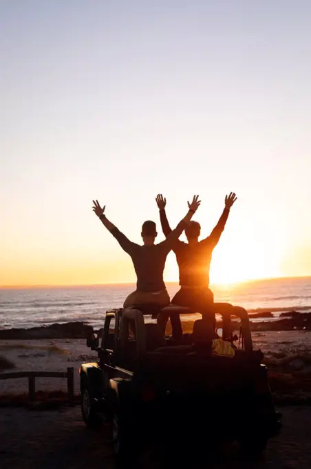 Rear view of caucasian gay male couple sitting on car roof raising arms at sunset by the sea. summer road trip and holiday in nature.