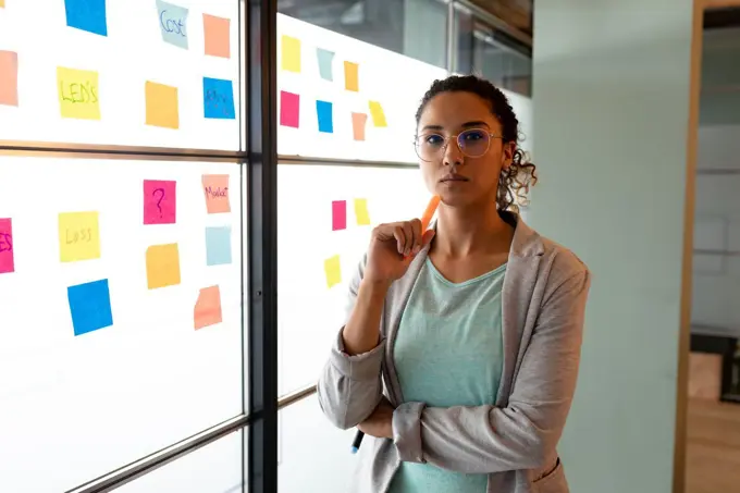 Portrait of confident biracial businesswoman in smart casuals by sticky notes on wall in office. creative business, modern office and business plan.