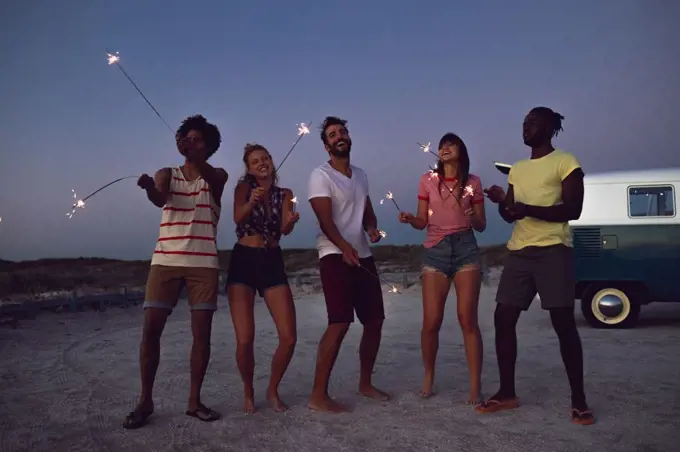 Front view of happy group of diverse friends playing with sparklers on the beach at dusk
