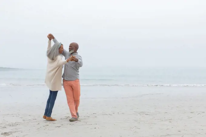 Front view of diverse happy senior couple smiling and dancing on the beach on cloudy day. Authentic Senior Retired Life Concept