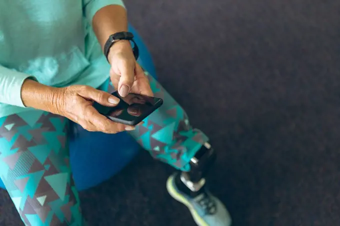 Close-up of disabled active senior Caucasian woman with leg amputee using mobile phone while sitting on exercise ball in fitness center. Strong active senior female amputee training and working out