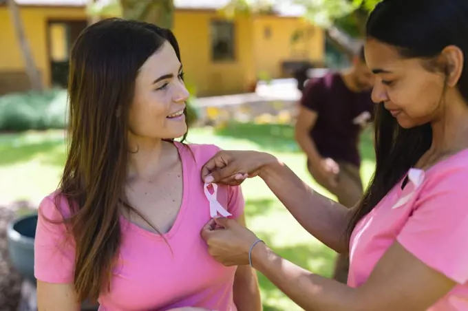 Biracial woman putting breast cancer awareness ribbon on t-shirt of caucasian female friend. breast cancer awareness campaign, friendship and unity concept.