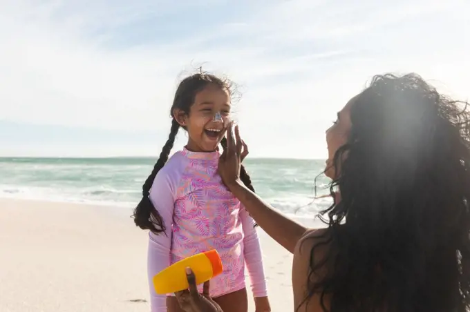 Cheerful biracial girl looking at mother applying sunblock lotion on her nose during sunny day. family and lifestyle and care.