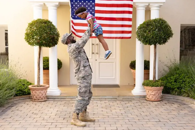 Happy african american military father holding aloft daughter in front of house with usa flag. family, bonding and patriotism, unaltered