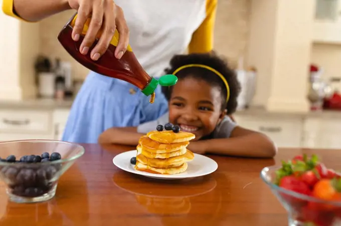 Midsection of mother putting maple syrup on pancake of african american daughter in kitchen at home. family, love and togetherness concept, unaltered.