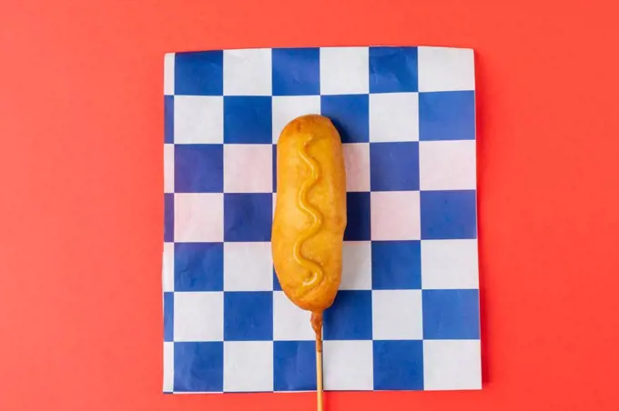 Directly above shot of corn dog with mustard sauce on checked pattern paper over red background. unaltered, unhealthy food, meat, sausage and fried food.