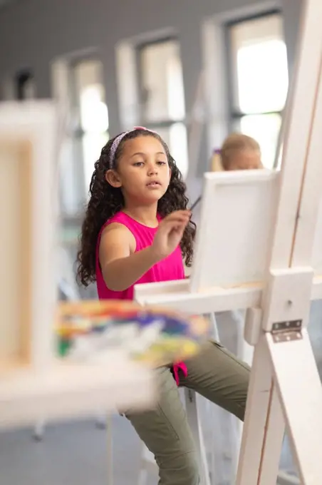 Cute biracial elementary schoolgirl painting on easel during drawing class in school. unaltered, childhood, education, art, painting, activity and back to school concept.