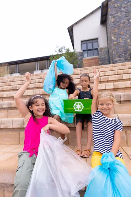 Happy multiracial elementary schoolgirls with garbage bags sitting on school steps. unaltered, sustainable lifestyle, education, cleaning, responsibility and recycling concept.