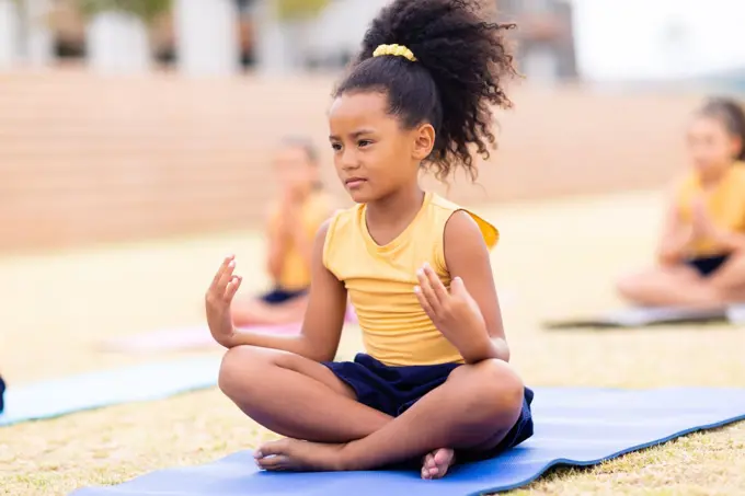 Biracial elementary schoolgirl gesturing while sitting on exercising mat at school ground. unaltered, childhood, education, activity, sports training, yoga and physical education concept.