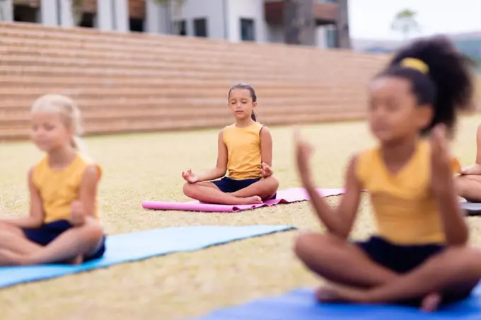 Multiracial elementary schoolgirls meditating while sitting on yoga mat at school ground. unaltered, childhood, education, activity, sports training, yoga and physical education concept.