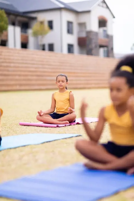 Multiracial elementary schoolgirls meditating while sitting on yoga mat against school. unaltered, childhood, education, activity, sports training, yoga and physical education concept.