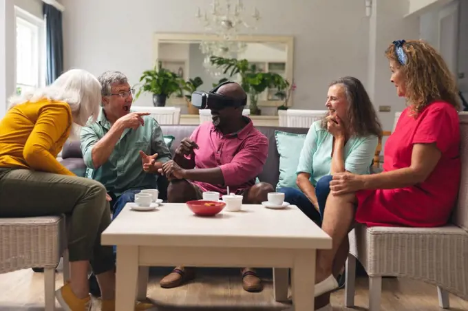 Happy multiracial senior man and women looking at male friend using vr headset at home. unaltered, lifestyle, social gathering, friendship, virtual reality, futuristic and modern technology.