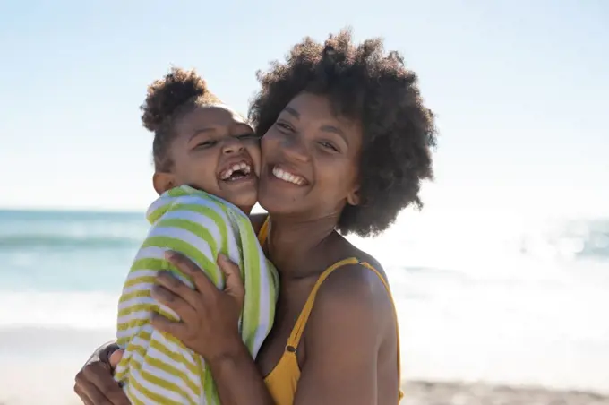 Portrait of cheerful african american mother and daughter with cheek to cheek at beach on sunny day. unaltered, family, lifestyle, togetherness, enjoyment and holiday concept.