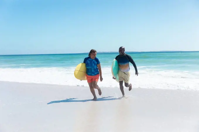 African american senior couple carrying surfboards running at beach against blue sky with copy space. unaltered, togetherness, active lifestyle, aquatic sport and holiday concept.