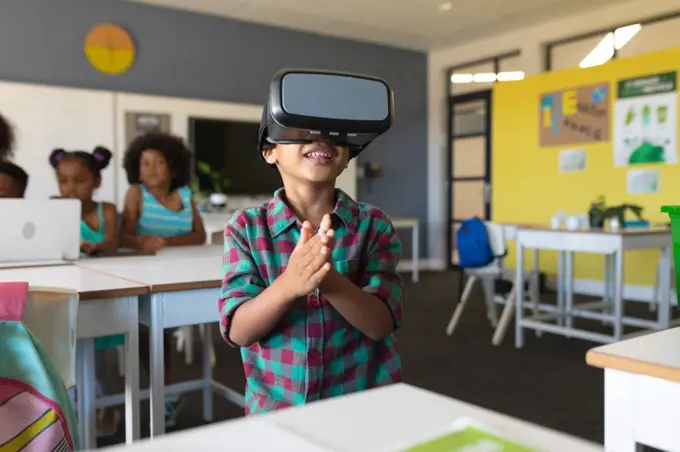 Smiling african american elementary schoolboy wearing vr glasses while standing in classroom. unaltered, education, virtual reality simulator, wireless technology and school concept.