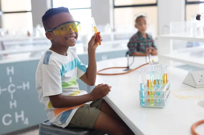 Portrait of smiling african american elementary schoolboy showing test tube during chemistry class. unaltered, education, learning, laboratory, stem, experiment, protection and school concept.