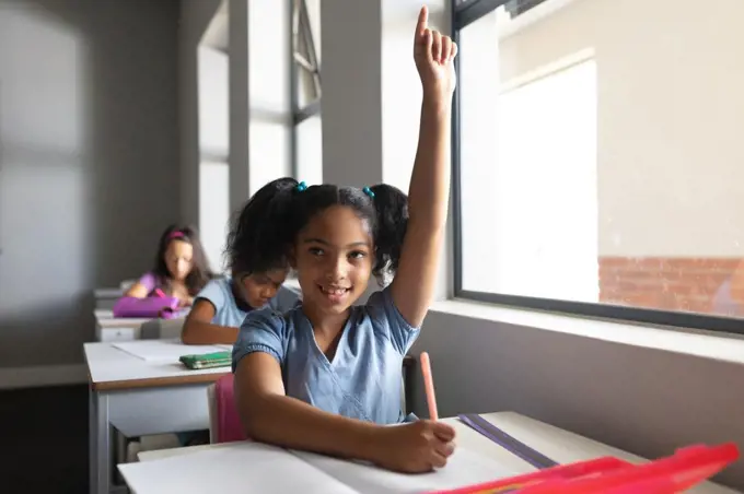 Smiling biracial elementary school girl with hand raised sitting at desk in classroom. unaltered, education, learning, childhood, intelligence and school concept.