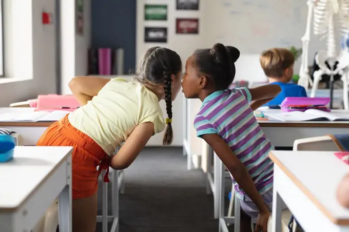 African american elementary girl whispering to caucasian female classmate at desk in classroom. unaltered, education, childhood, gossip, secret, sharing, science, stem and school concept.