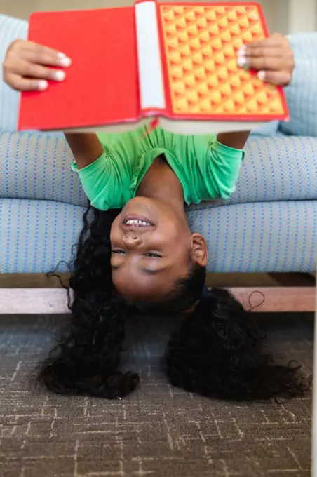 Smiling african american elementary girl reading book while lying upside down on couch in school. unaltered, education, childhood, relaxation, reading, studying and school concept.