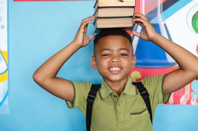 Portrait of smiling african american elementary schoolboy stacking books on head against wall. unaltered, education, childhood and school concept.