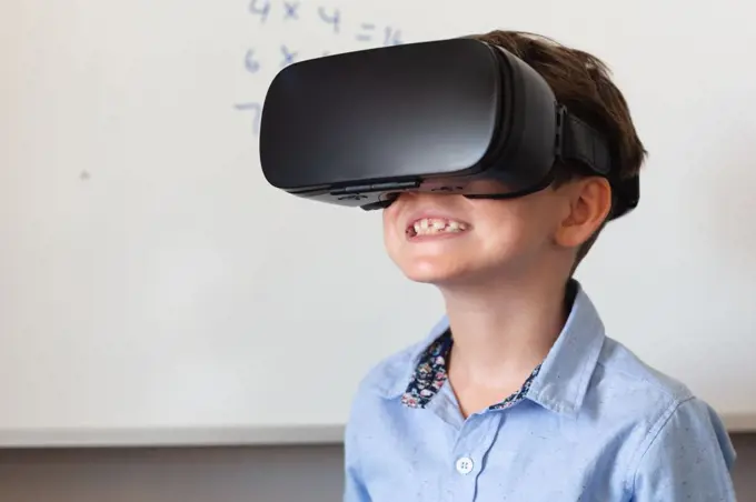 Smiling caucasian elementary schoolboy wearing vr glasses against whiteboard in classroom. unaltered, education, virtual reality simulator, technology and school concept.