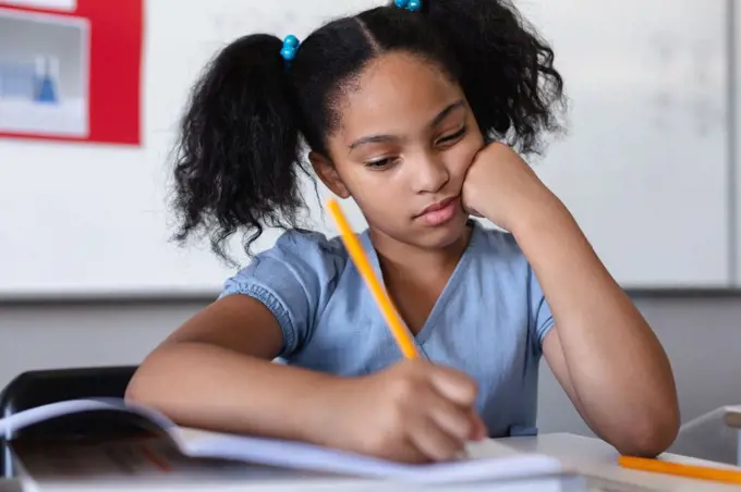 Biracial elementary schoolgirl writing on book at desk in classroom. unaltered, education, learning, studying, concentration and school concept.