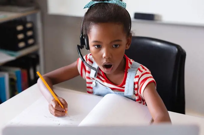 African american elementary schoolgirl with mouth open looking at laptop while studying at desk. unaltered, education, listening, studying, concentration, wireless technology and school concept.