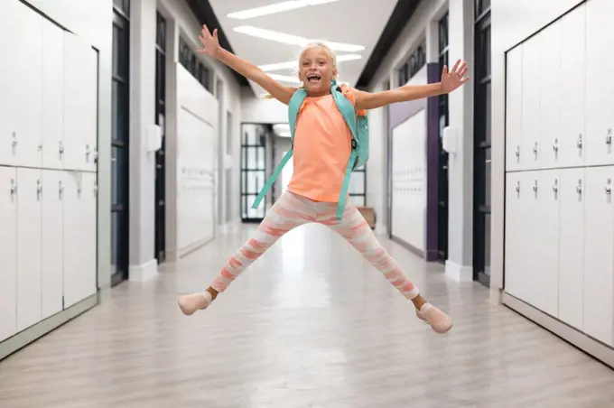 Full length of caucasian elementary schoolgirl with arms outstretched jumping in corridor. unaltered, childhood, education, happiness, playful and school concept.