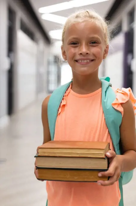 Portrait of smiling caucasian elementary schoolgirl holding books while standing in corridor. unaltered, childhood, education, happiness, learning and school concept.