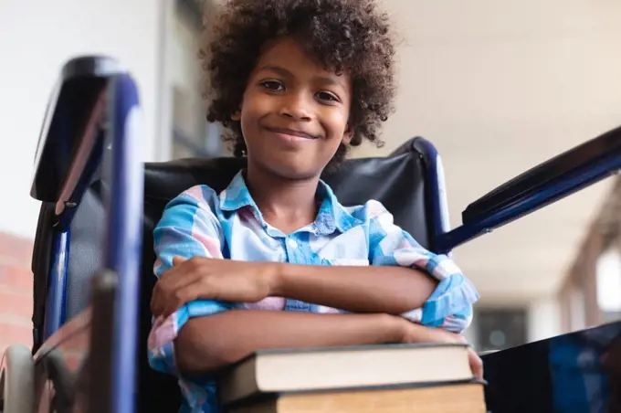 Low angle portrait of smiling african american elementary schoolboy with books sitting on wheelchair. unaltered, education, childhood, physical disability, and school concept.