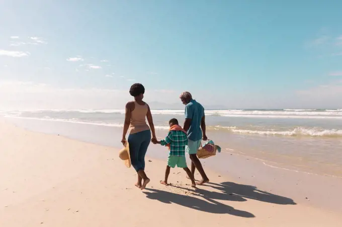 African american grandparents holding boy's hands and walking at sandy beach against sky in summer. nature, copy space, unaltered, family, love, togetherness, childhood, enjoyment, holiday concept.